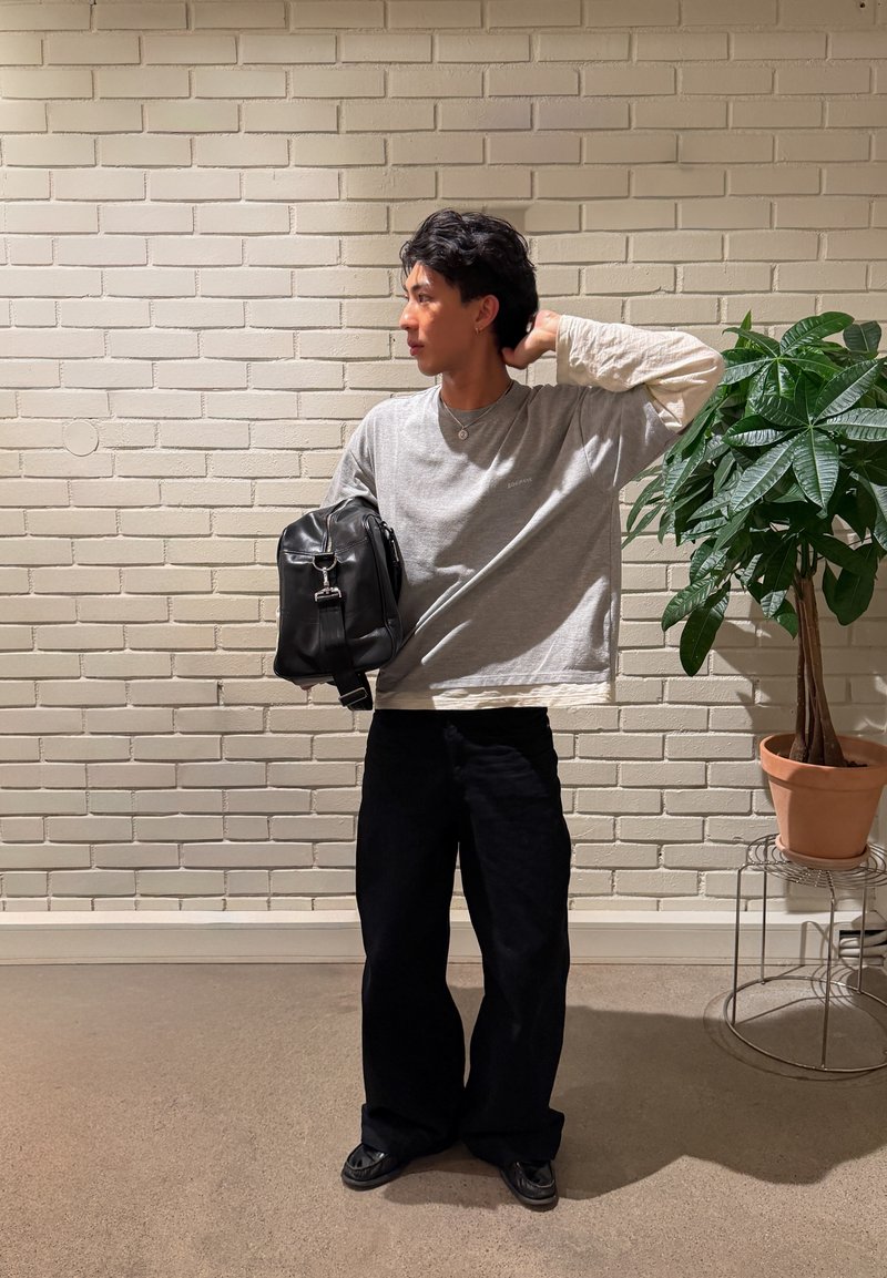 Jeune homme debout contre un mur de briques blanches, portant une chemise grise à manches longues, un pantalon noir, tenant un sac noir, avec une plante en pot à proximité.