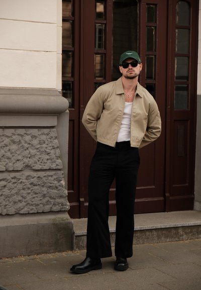 Chaqueta beige con cremallera sobre una camiseta blanca sin mangas, combinada con pantalones negros y botas negras; usando gafas de sol oscuras y una gorra verde.