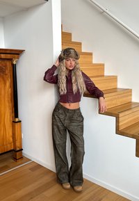 Burgundy läderkort jacka, olivgröna läderbyxor med vida ben, brun pälsmössa, står vid en trätrappa mot en vit vägg.