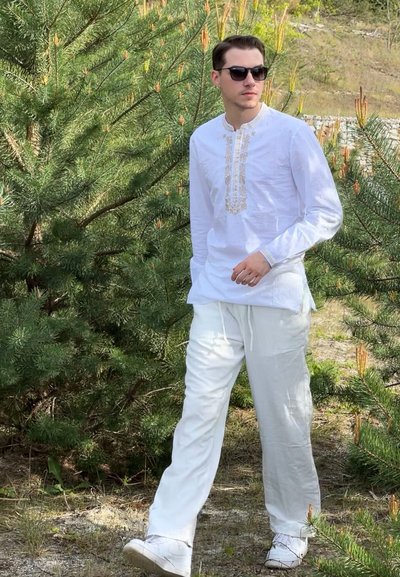 Camisa de lino blanca de manga larga con cuello bordado, combinada con pantalones blancos y zapatillas, en un fondo de pinos verdes.