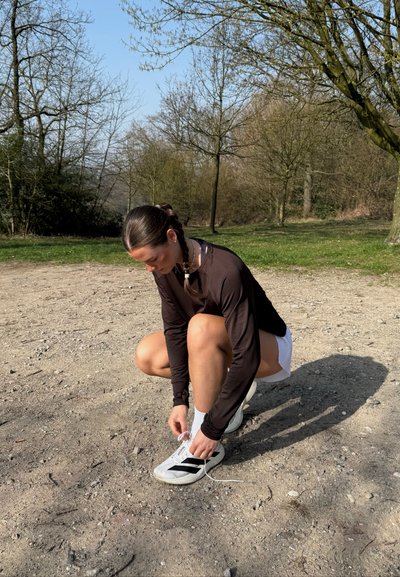 Mujer con el cabello trenzado, vestida con una camiseta negra de manga larga y pantalones cortos blancos, atándose los tacos de fútbol blancos en un camino de grava en un parque.