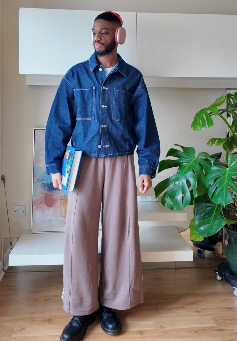 Homme portant une veste en jean, un pantalon large beige et un casque rose, tenant un livre, debout près d'une grande plante verte en pot à l'intérieur.