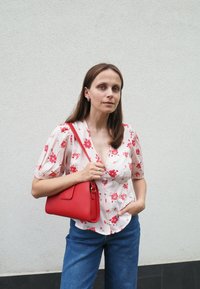 Raudona odinė rankinė, keturkampio formos ir su trumpa petneša. dėvima su gėlių rašto trumpomis rankovėmis bliuskėmis ir mėlynomis džinsomis.