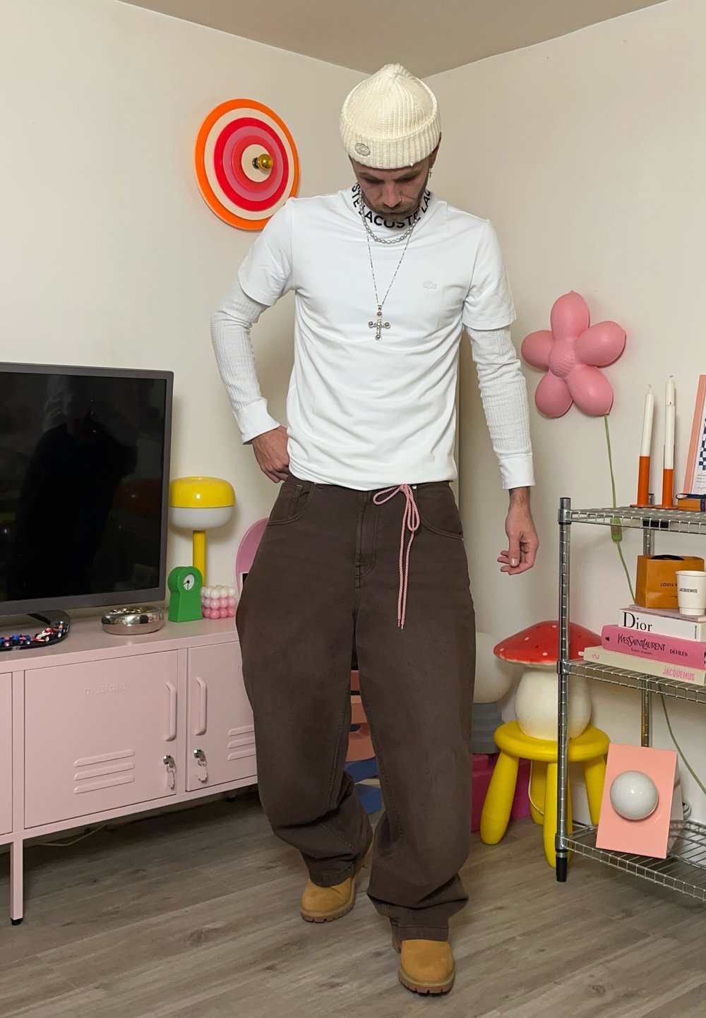 Camisa branca de manga longa com detalhes cinzentos, calças largas castanhas com um cordão rosa, e botas bege. Decoração de parede redonda e prateleiras coloridas ao fundo.