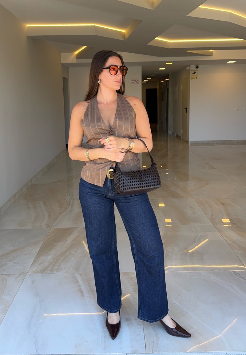 Femme portant un haut marron sans manches, un jean bleu foncé, des chaussures pointues marron, des lunettes de soleil orange, et tenant un sac à main noir dans un couloir moderne.