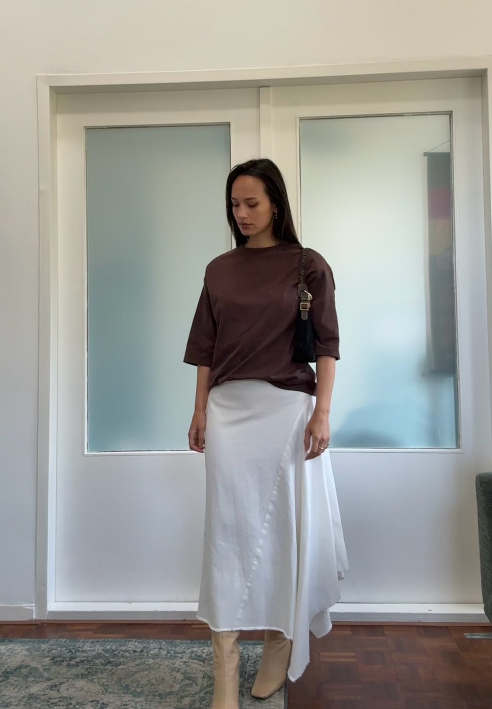 Camisetta marrone a maniche corte abbinata a una gonna bianca asimmetrica. La gonna è lunga fino al pavimento e ha una texture fluida. Stivali alla caviglia beige.