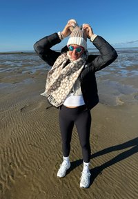 Personne portant une doudoune noire, un chapeau en tricot beige et une écharpe grise à motif tacheté, des leggins noirs et des baskets blanches. Cadre ensoleillé sur la plage.