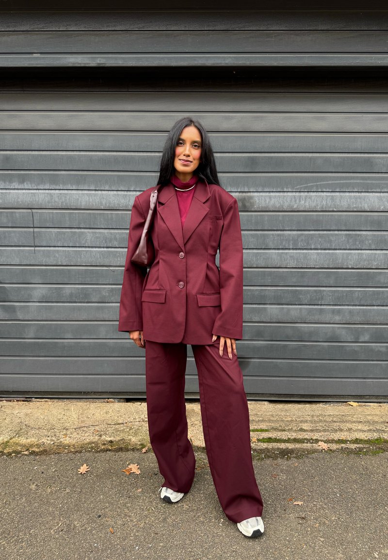 Femeie în costum larg bordo și adidași, stând în fața unei uși metalice gri de garaj, în aer liber.