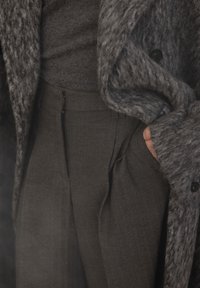 Personne portant un manteau gris texturé et un pantalon gris à motif chevrons taille haute, main posée de manière décontractée dans la poche.