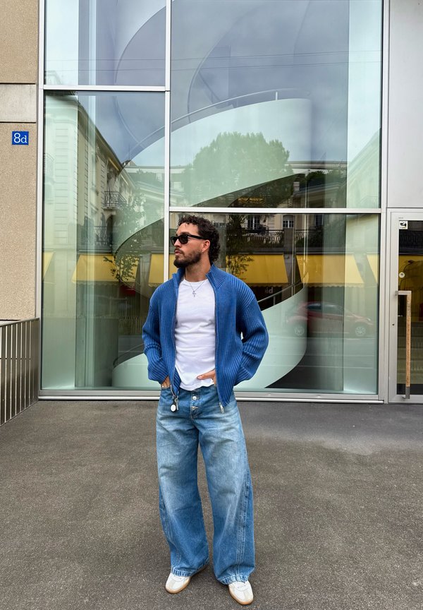 Hombre con gafas de sol, cárdigan azul, camisa blanca y vaqueros anchos de mezclilla de pie frente a una pared de cristal con una escalera de caracol en su interior.
