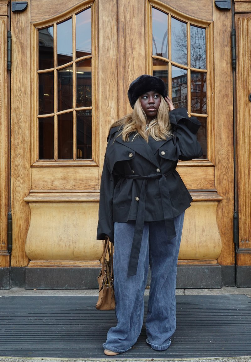 Trench-coat noir avec des boutons dorés porté sur un haut gris. Pantalon large en denim et chapeau en fourrure noire. Sac marron à la main, posé devant des portes en bois.