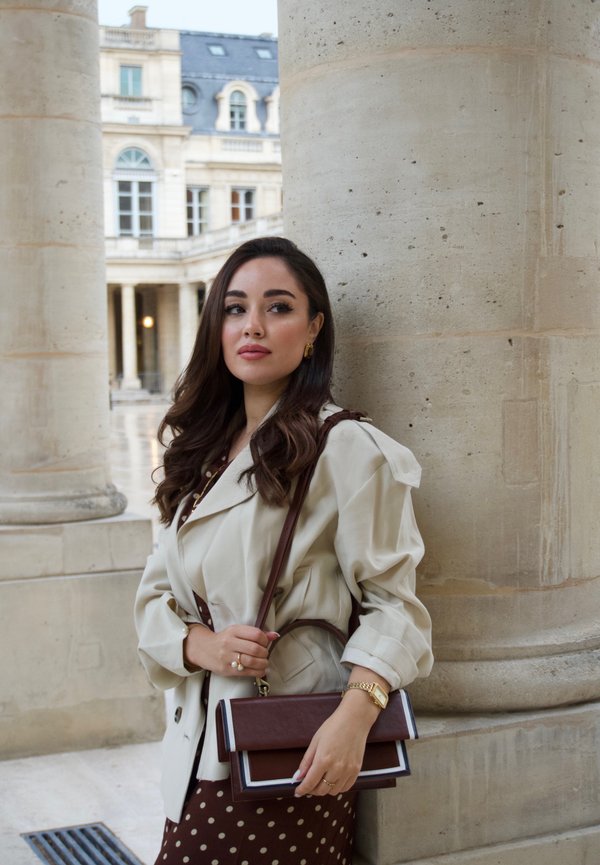Beige Trenchcoat mit Puffärmeln, braunes Kleid mit Punkten, braune und cremefarbene Handtasche mit Klappe, goldener Schmuck und welliges braunes Haar.