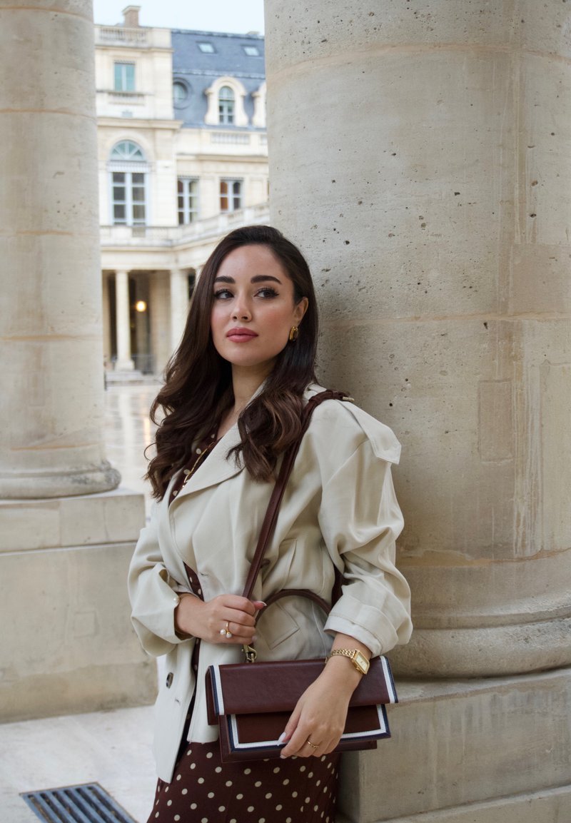 Beige Trenchcoat mit Puffärmeln, braunes Kleid mit Punkten, braune und cremefarbene Handtasche mit Klappe, goldener Schmuck und welliges braunes Haar.