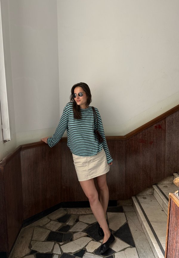 Femme aux longs cheveux foncés portant des lunettes de soleil, un chemisier rayé bleu et noir à manches longues, une jupe blanche et des chaussures noires, appuyée sur une rambarde près d'escaliers.