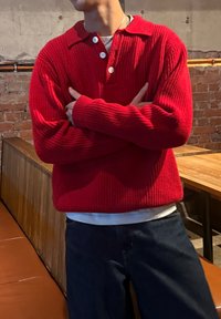 Pull en maille rouge avec texture côtelée, col à revers, patte à trois boutons et coupe ample, associé à un jean bleu foncé dans un décor rustique.