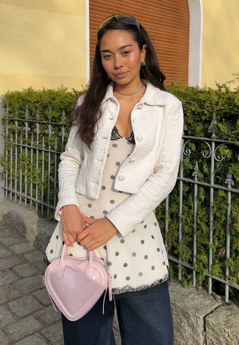 Młoda kobieta stojąca na zewnątrz przy metalowym ogrodzeniu, ubrana w białą, teksturowaną kurtkę, bluzkę w grochy, ciemne spodnie i trzymająca różową torebkę w kształcie serca.
