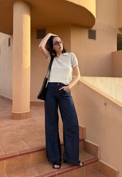 Camisa polo de manga corta beige, jeans de pierna ancha azul oscuro, bolso negro y sandalias de tiras negras; de pie en unas escaleras con una pared texturizada detrás.