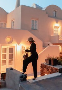 Una donna in un completo nero e un cappello sale su un bordo di pietra. Sullo sfondo si scorge un edificio in stucco bianco illuminato delicatamente con finestre.