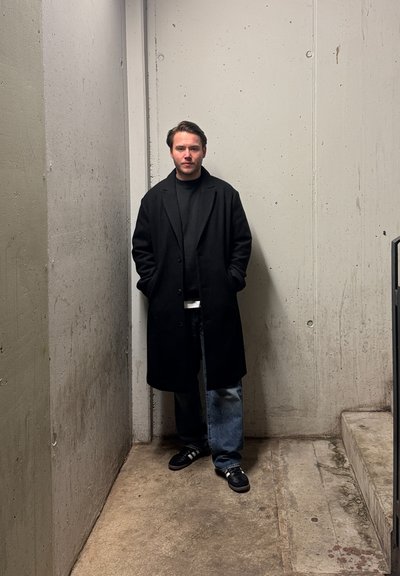 Abrigo largo negro sobre un suéter negro, combinado con jeans azules y zapatillas negras. El escenario es un espacio de concreto texturizado con escaleras.