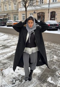 Manteau à rayures noires à double boutonnage porté sur un haut blanc transparent à volants et une chemise foncée côtelée, associé à un pantalon gris et de grandes bottes noires.