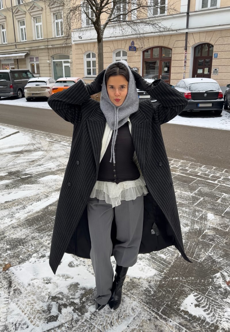 Manteau croisé à rayures noires porté sur un haut blanc transparent à volants et une chemise côtelée foncée, assorti à un pantalon gris et de grandes bottes noires.