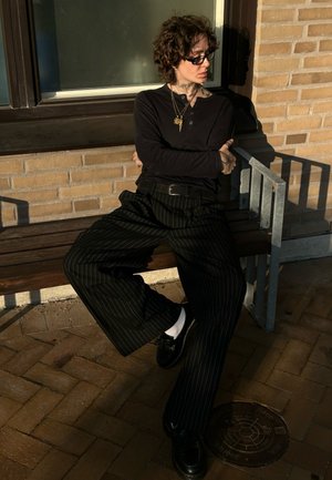 Svart långärmad skjorta och randiga vida byxor, bärs med svarta skor och vita strumpor, sitter på en träbänk, solljus som kastar skuggor.