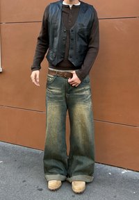 Chaleco de cuero negro sobre un suéter marrón de canalé, combinado con unos vaqueros anchos de color azul claro. El conjunto incluye un cinturón marrón y botas beige.