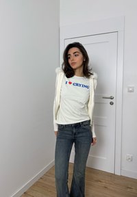 Fehér hosszú ujjú ing kék és piros "I ❤️ CRYING" felirattal, fehér szőrmés kardigán alatt, bővülő kék farmerral párosítva.