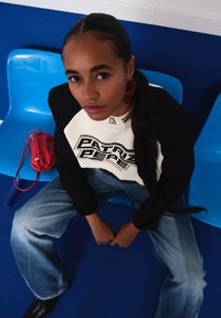 Jovem mulher com trança longa, vestindo um sweatshirt preto e branco e jeans largos, senta-se em uma cadeira de plástico azul com uma pequena bolsa vermelha ao seu lado.