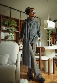 Grå oversized genser med en drapert skjerf, paret med løse brune bukser. Plassert i et moderne interiør med planter og minimalistisk dekor.