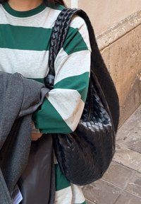 Personne portant un chemise à manches longues rayée vert et blanc, portant un sac en cuir tressé noir et tenant un manteau gris contre un mur en pierre.