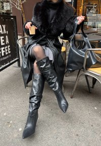 Personne portant des bottes en cuir noires, un manteau à col en fourrure noire, tenant une tasse de café et un grand sac en cuir noir dans un café en plein air.
