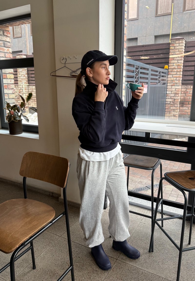 Person in lässiger schwarzer Kappe, schwarzem Sweatshirt, grauen Jogginghosen und Stiefeln, die einen grünen Becher hält und in einem modernen Café am Fenster steht.