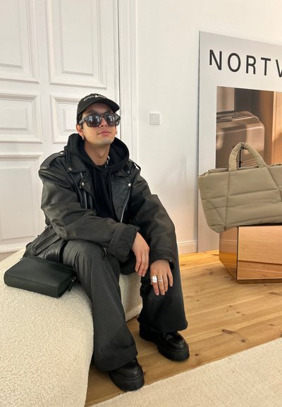 Chaqueta de cuero negra, sudadera negra, pantalones grises y zapatos negros gruesos. Los accesorios incluyen grandes gafas de sol negras y un bolso de mano verde.