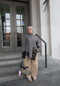 Femme portant une écharpe grise, un pull boutonné, un pantalon large beige avec un détail en dentelle noire, des gants noirs, tenant un sac à main à motif avec un porte-clés en peluche rose.