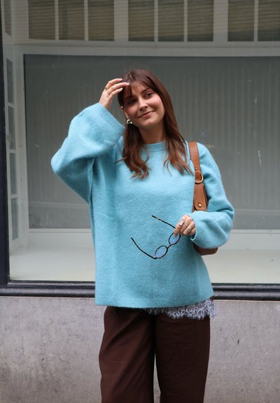 Mujer con suéter azul sosteniendo gafas en una mano, tocándose el cabello con la otra, de pie al aire libre frente a una ventana.