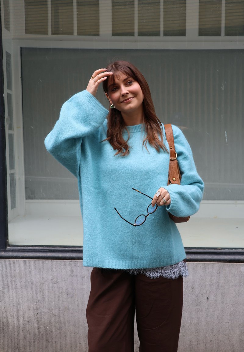 Vrouw in blauwe trui houdt een bril in één hand vast, raakt met de andere haar haar aan, staat buiten tegen een raamachtergrond.