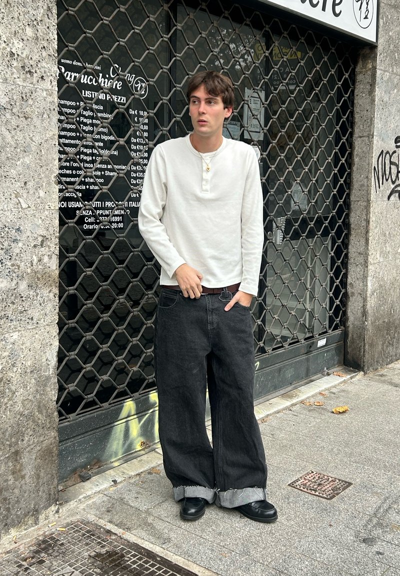 Chemise henley blanche à manches longues, jean noir à jambes larges avec ourlets retroussés, et chaussures noires. En arrière-plan, une grille en métal et une enseigne de magasin.