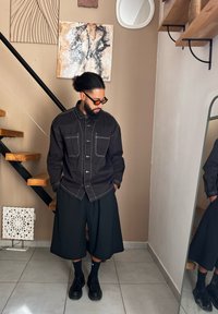 Μαύρο πουκάμισο με κουμπιά, δύο μπροστινές τσέπες, λευκές ραφές, συνδυασμένο με φαρδιά μαύρη βερμούδα. Μαύρα παπούτσια και κάλτσες ολοκληρώνουν την εμφάνιση.