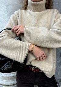 Beige Rollkragenpullover mit gerippten Bündchen, kombiniert mit einer schwarzen geflochtenen Handtasche und goldenen Armreifen. Ein brauer Gürtel ist auf dunklen Hosen sichtbar.