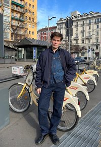 Νέος άνθρωπος που φοράει ένα σκούρο μπλε τζιν μπουφάν πάνω από μαύρο bomber μπουφάν, σκούρα μπλε τζιν και μαύρα παπούτσια, με κίτρινες μοτοσικλέτες στο παρασκήνιο.