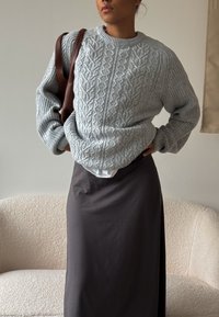 Grijze kabelgebreide trui met een gestructureerd ontwerp, gecombineerd met een lange donkere rok. De trui heeft een ronde halslijn en geribde manchetten. Bruine tasaccenten.