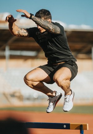 Atleta con brazos tatuados, vistiendo pantalones cortos negros, salta sobre una valla en una pista al aire libre bajo un cielo despejado.
