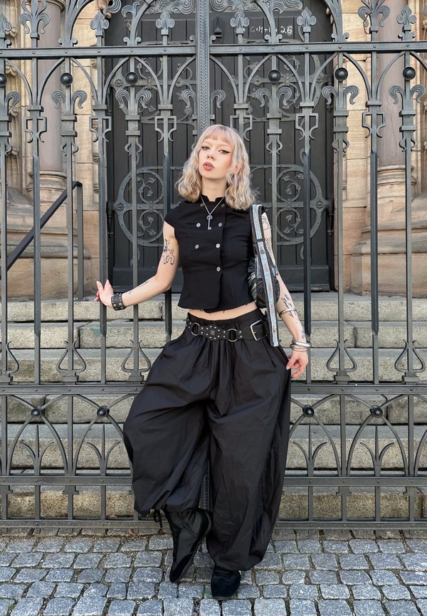 Personne aux cheveux blonds mi-longs portant un haut court noir et un pantalon large, debout devant une porte en fer ornée et des marches en pierre.