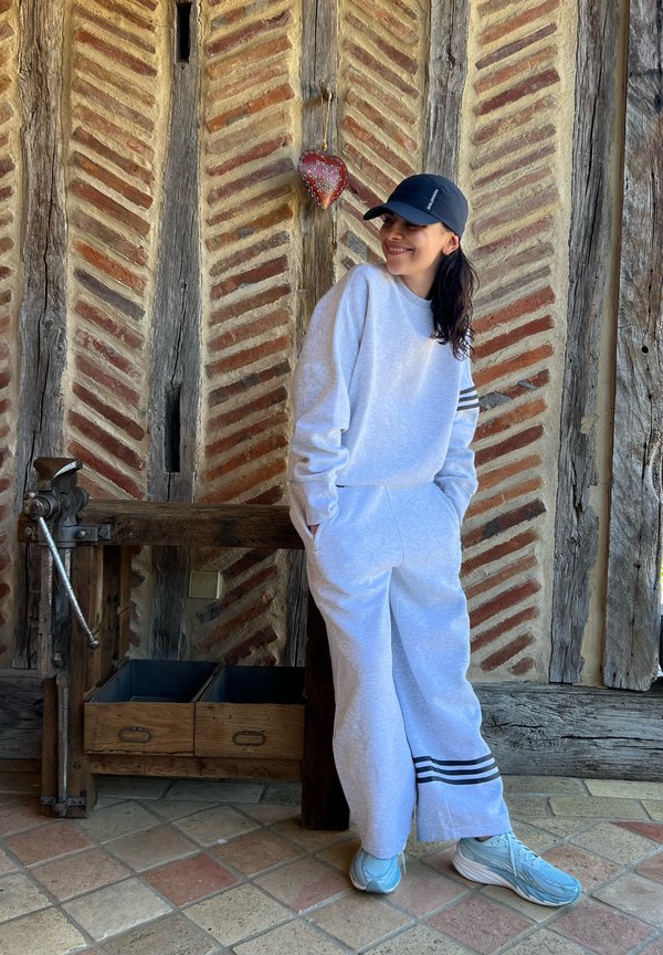 Femme en survêtement gris clair et casquette noire debout contre un mur en brique et bois à motifs avec une petite décoration de cœur rouge suspendue au-dessus.