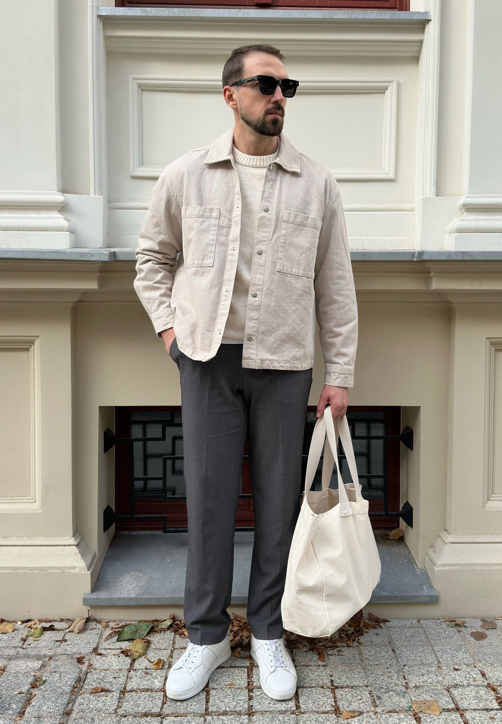 Lys beige knap-opp jakke over en cremefarvet sweater, grå bukser, hvide sneakers og en stor cremefarvet tote bag; stående mod en stenmur.