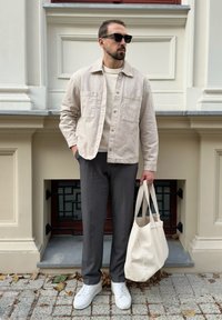 Lys beige knap-opp jakke over en cremefarvet sweater, grå bukser, hvide sneakers og en stor cremefarvet tote bag; stående mod en stenmur.