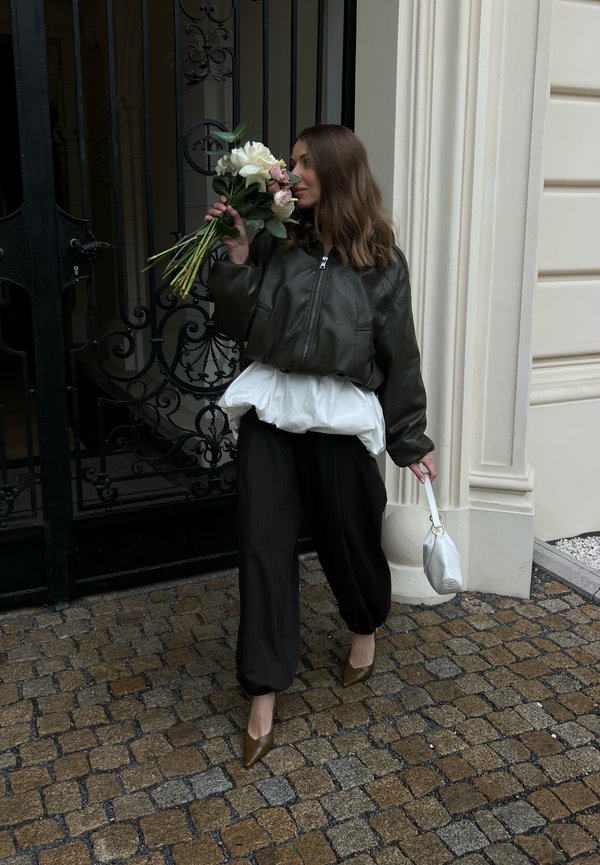 Femme marchant sur une rue pavée tenant un bouquet près d'une grille en fer forgé, portant un pantalon noir, une veste sombre et un sac à main blanc.