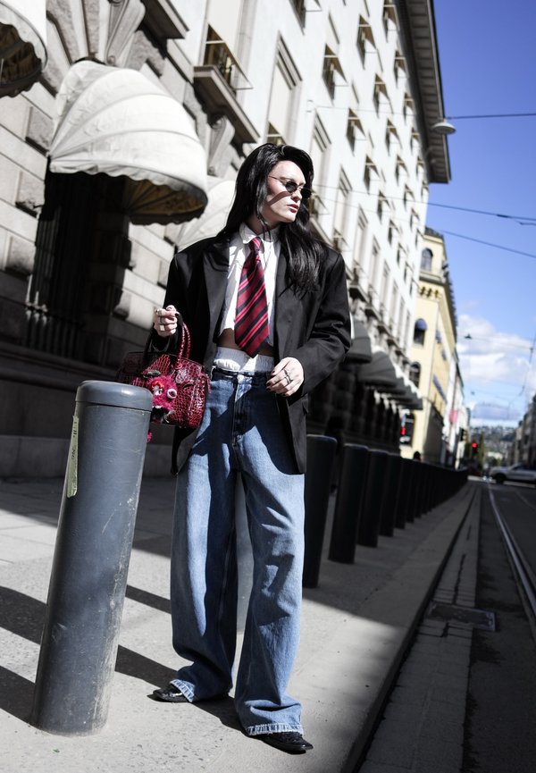 Person mit langen schwarzen Haaren, die eine Sonnenbrille trägt, übergroßen schwarzen Blazer, rote gestreifte Krawatte, weite Jeans und eine rote strukturierte Handtasche auf der Stadtstraße hält.