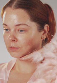 Nærbilde av en person med lys hud og rødt hår, som har på seg et rosa satengplagg og fjærpynt, mens de forsiktig berører haken sin.